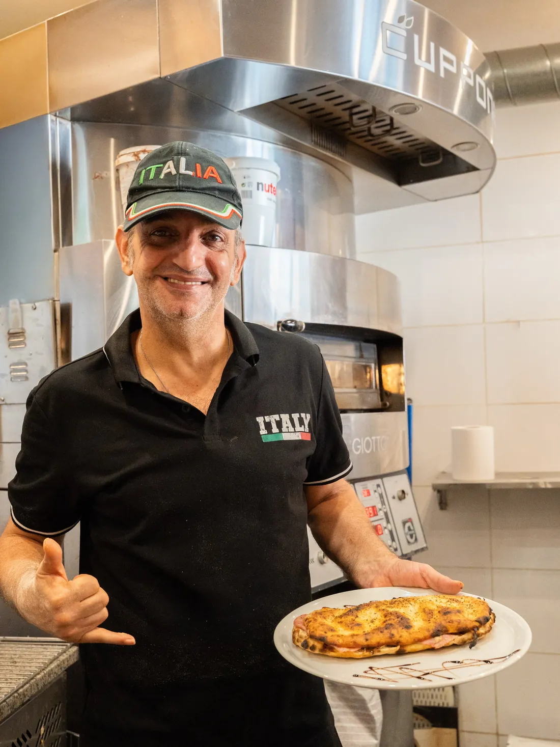 Gio, casquette Italia, montre fièrement une pizza devant le four
