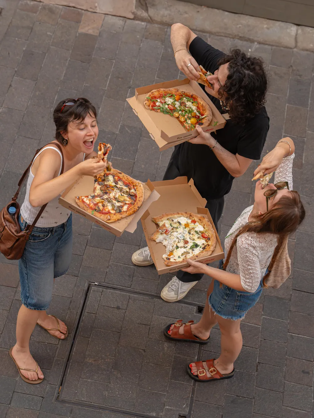 Trois amis mangent leurs pizzas debout, vus d’en haut